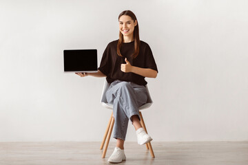 Poster - Smiling young woman sitting on chair, showing laptop with empty screen, gesturing thumb up, mockup for website. Millennial lady with portable computer recommending your product ad