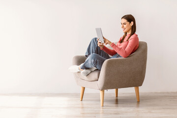 Wall Mural - Happy millennial lady using tablet pc, studying or working remotely, having online conference, sitting in armchair against white studio wall, copy space. Young woman with touch pad checking new app