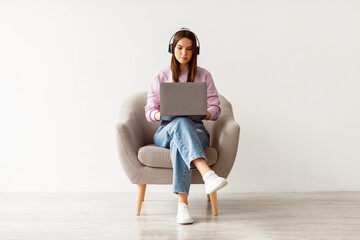 Poster - Young Caucasian woman having video call, using laptop computer, sitting in armchair, wearing wireless headphones against white studio wall, full length. Distance communication concept