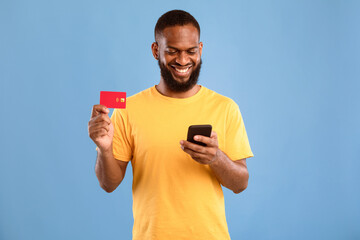 Aufkleber - Positive African American guy holding credit card and using smartphone, shopping online on blue studio background. Millennial man making contactless payment. Web banking service, cashback concept