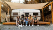 © Prostock-studio - Group of cheery young diverse people with their cute dog sitting near motorhome, having fun camping trip in autumn. Multiethnic millennial friends relaxing together on weekend in countryside