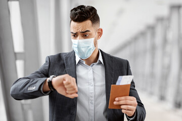 Wall Mural - Worried Arab Man In Medical Mask Looking At Wristwatch, Getting Late For Flight, Anxious Middle Eastern Man Holding Passport And Tickets, Hurry To Boarding Gate, Travelling During Pandemic, Closeup