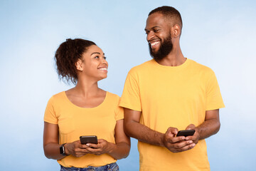 Poster - Millennial people communication. African American woman and man texting on mobile phones, standing side by side on blue studio background. Couple using modern gadgets