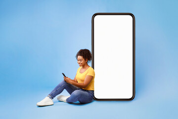 Poster - Cheery African American lady sitting near big cellphone with empty white screen, using mobile device, checking new cool app on blue studio background, mockup for website or application design