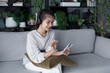 © Wanda - Asian woman sits on sofa listening to music with smartphone in living room