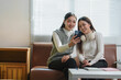 © crizzystudio - Two young asian women, likely students or colleagues, are taking a selfie with a smartphone while sitting on a couch in a modern, light-filled room, possibly a study area or office break room