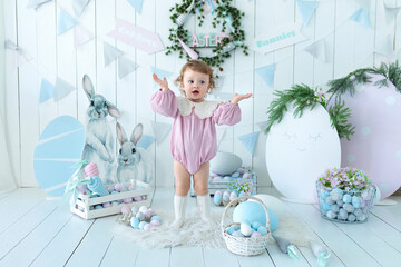 Wall Mural - Cute smile baby girl with curly hair and in pink bodysuit in easter decorations at home. Happy child with easter eggs and decorative figurine bunnies. Little girl in child room in the spring style. 