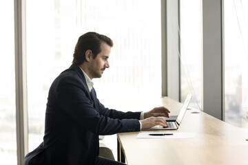 Wall Mural - Young 35s businessman in suit typing on his laptop in workspace, looks focused, lead correspondence to client, send e-mail, solve business remotely, prepare report, work on presentation. Trading, tech