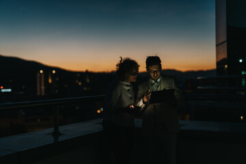 Wall Mural - Two business people collaborate on a project at night using a digital tablet. The backdrop of a city skyline at dusk adds to the atmosphere of creativity and focus.