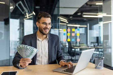 Wall Mural - Successful joyful businessman working inside office with laptop. Man in business suit holding money cash American dollars in hands. Online investor financier satisfied with results of achievement.