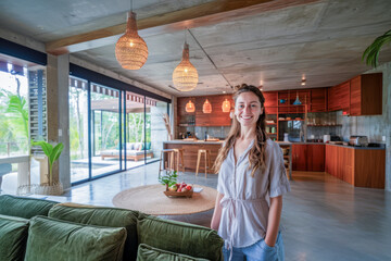 Wall Mural - Happy woman in a spacious, modern open-plan kitchen and living area with tropical decor, natural lighting, and warm wooden elements