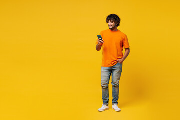 Wall Mural - Full body young smiling happy Indian man he wears orange t-shirt casual clothes hold in hand use mobile cell phone chat online isolated on plain yellow background studio portrait. Lifestyle concept.