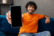 © ViDi Studio - Young Indian man wear orange casual clothes sits on blue sofa couch hold use blank screen mobile cell phone stay home hotel flat rest relax spend free spare time in living room indoor. Lounge concept.