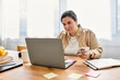 © Bliss - A young plus sized woman works on her laptop at a wooden table while holding her phone.