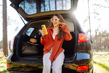 Wall Mural - Beautiful woman sits in the trunk of a car outdoor and uses a smartphone. Travel, weekend, nature, relax and lifestyle concept. Blogging, communication.