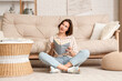 © Pixel-Shot - Young pretty woman reading book and sitting on floor near sofa in living room