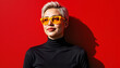 © Alina - Confident young woman with short hair wearing bold yellow sunglasses against red wall
