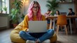 © IgorImageDoc - young woman pink hair sitting yellow bean bag chair living room she wearing yellow sweater blue jeans white sneakers she has laptop her lap looking directly camera smile her face background two