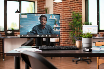 Wall Mural - Empty brick wall room equipped with desktop showing black businessman conducting virtual meeting. African american manager on videoconference call displayed on computer screen in office with no people