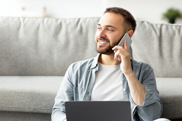 Poster - Full length of happy young guy using laptop and mobile phone at home, having freelance job. Millennial man chatting with friends, having phone call during online business meeting