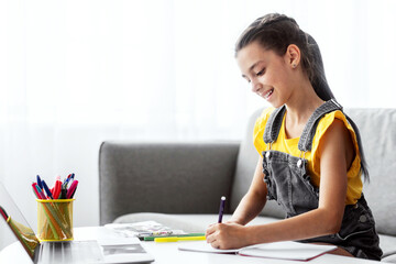 Poster - Portrait of smiling schoolgirl sitting on couch at desk with pc, writing essay or drawing picture in paper notebook. Pupil studying or taking notes in her diary, making checklist, free copy space