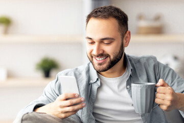 Poster - Positive millennial guy holding modern smartphone and cup of coffee indoors, chatting with friends, sending email, browsing internet or watching entertainment video. Online life concept