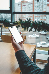 Aufkleber - A person holding a smartphone with a blank white screen near a wooden table by a large window. The view outside shows urban buildings and cars with festive decorations on the window