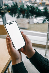 Aufkleber - A person holding a smartphone with a blank white screen near a wooden table by a large window. The view outside shows urban buildings and cars with festive decorations on the window