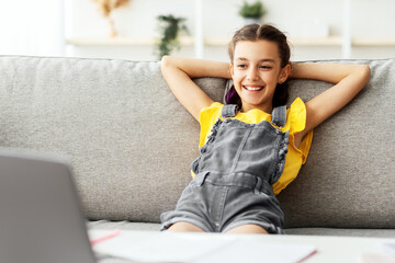 Poster - Rest Concept. Smiling female teenager sitting on comfortable couch, leaning back on sofa and looking at laptop screen, watching movie or video, taking break from studying online at home