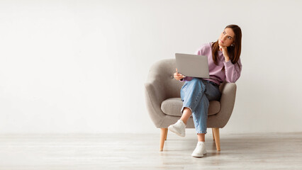 Poster - Deadline stress concept. Frustrated young woman sitting in armchair with laptop computer, looking away deep in thought, feeling tired after long working day against white studio wall, full length
