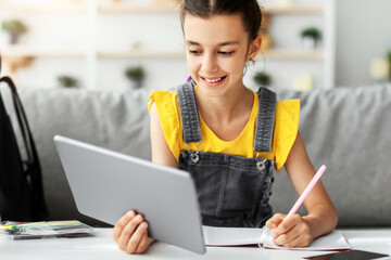 Poster - Portrait of smiling junior school kid girl holding digital tablet studying online during virtual class learning at home, doing homework and taking notes in her copybook or having video call with tutor