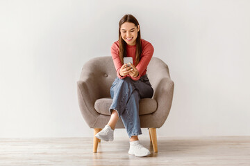 Poster - Happy Caucasian woman using cellphone, chatting on internet, working or studying online, sitting in armchair against white studio wall, full length. Cheery young lady watching video on mobile phone