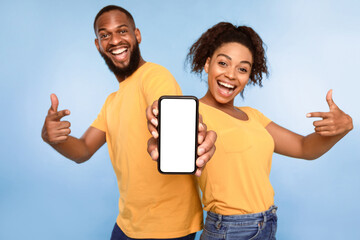 Poster - Mobile application ad. Excited African American couple pointing at cellphone with blank screen, recommending new app on blue studio background, selective focus. Mockup for website