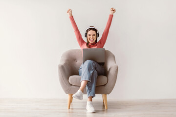 Poster - Full length of excited young lady with laptop and headphones raising her arms up, winning online casino bet, shopping on sale, reaching goal, achieving success, sitting in armchair against white wall