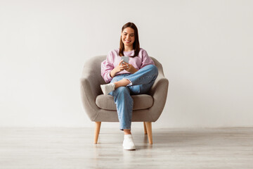 Poster - Beautiful young woman sitting in armchair, looking at mobile phone screen, using new cool application against white studio wall, full length. Joyful lady using smartphone for work or entertainment