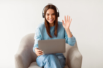 Poster - Distant virtual communication concept. Pretty young lady in headphones waving at laptop screen, sitting in armchair, making video call, chatting online on white studio background