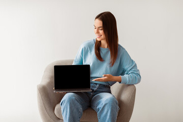 Poster - Smiling millennial woman in casual clothes showing laptop with empty screen, sitting in armchair on white studio background, mockup for website. Young lady presenting portable computer with template