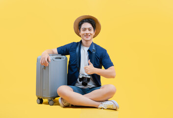 Wall Mural - Full body image of a young Asian man holding a suitcase and wearing a travel camera posing on a yellow background