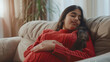 © Shivani - Indian teens girl feeling hungry while holding her belly on the couch in a Living room, she is closing her eyes, wearing red sweater
