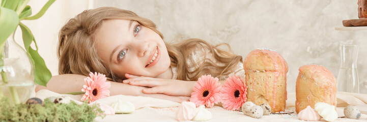 Wall Mural - A girl with long hair in a light dress is sitting at the Easter table with cakes, spring flowers and quail eggs. Happy Easter celebration.