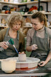 © Irina_Evva - Mentor teaches pottery fundamentals, demonstrating sculpting skills on an electric wheel Active aging concept, retired woman explores pottery as a new passion using a spinning wheel