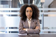 © Tetiana - Portrait of a young serious African American business woman in a suit and glasses crossing her arms on her chest, sitting at the desk in the office and looking confidently at the camera