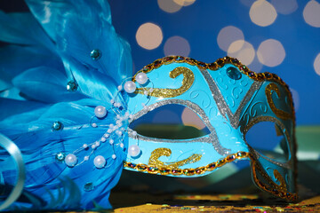 Wall Mural - Beautiful carnival mask and confetti on table against blue background with blurred lights, closeup