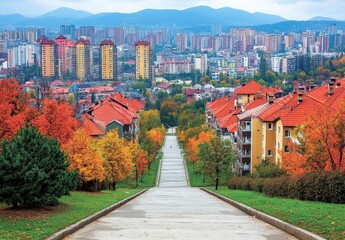 Canvas Print - A view of a city from an elevated pathway, featuring colorful trees and buildings in various shades. The pathway leads toward the city, lined with