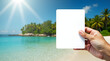 © Anastasia - A person holding a blank card in front of a sunny beach with clear blue water and palm trees creating a serene vacation atmosphere