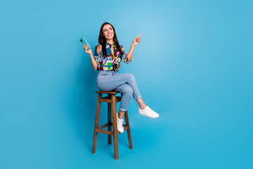 Poster - Full size photo of positive girl dressed blouse sit on chair hold smartphone look directing empty space isolated on blue color background
