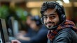 © Yuliia - Young South Asian male customer service representative wearing headset and casual attire smiles while working at computer in modern office environment.
