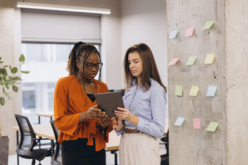 Wall Mural - Professional women reviewing project data on tablet, collaborating near colorful sticky notes wall in contemporary workspace