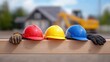 © Moopingz - Three colorful hard hats and work gloves rest on a wooden beam at a construction site, with a house and excavator in the background.