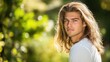 © The 2R Artificiality - Handsome young man with long wavy hair in a sunlit outdoor setting surrounded by lush green foliage wearing a casual white shirt.